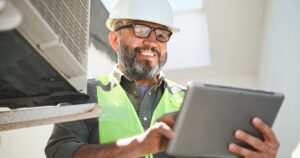 middle aged man with salt and pepper beard standing next to air conditioning field service equipment and holding a tablet.